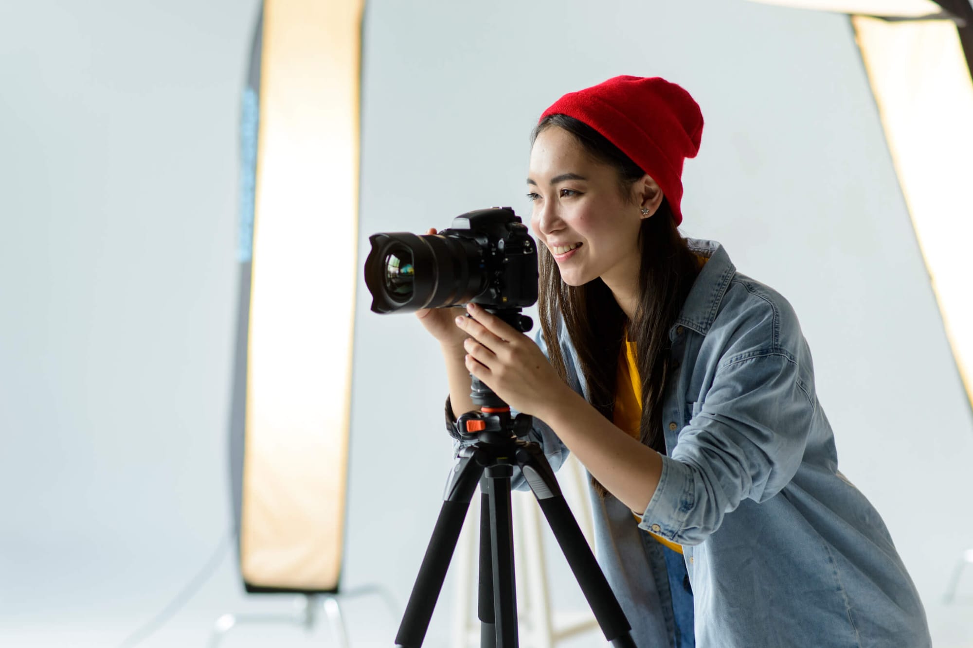 A young woman in a red beanie and denim jacket works with a professional camera on a tripod, wearing a casual outfit.