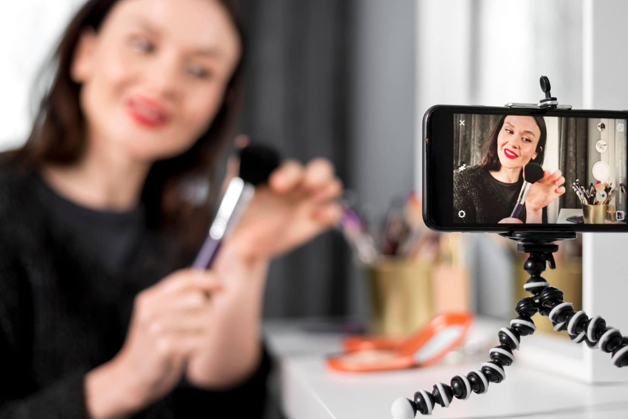 A woman records a makeup tutorial on her phone, holding a brush, with the phone mounted on a flexible tripod in focus.