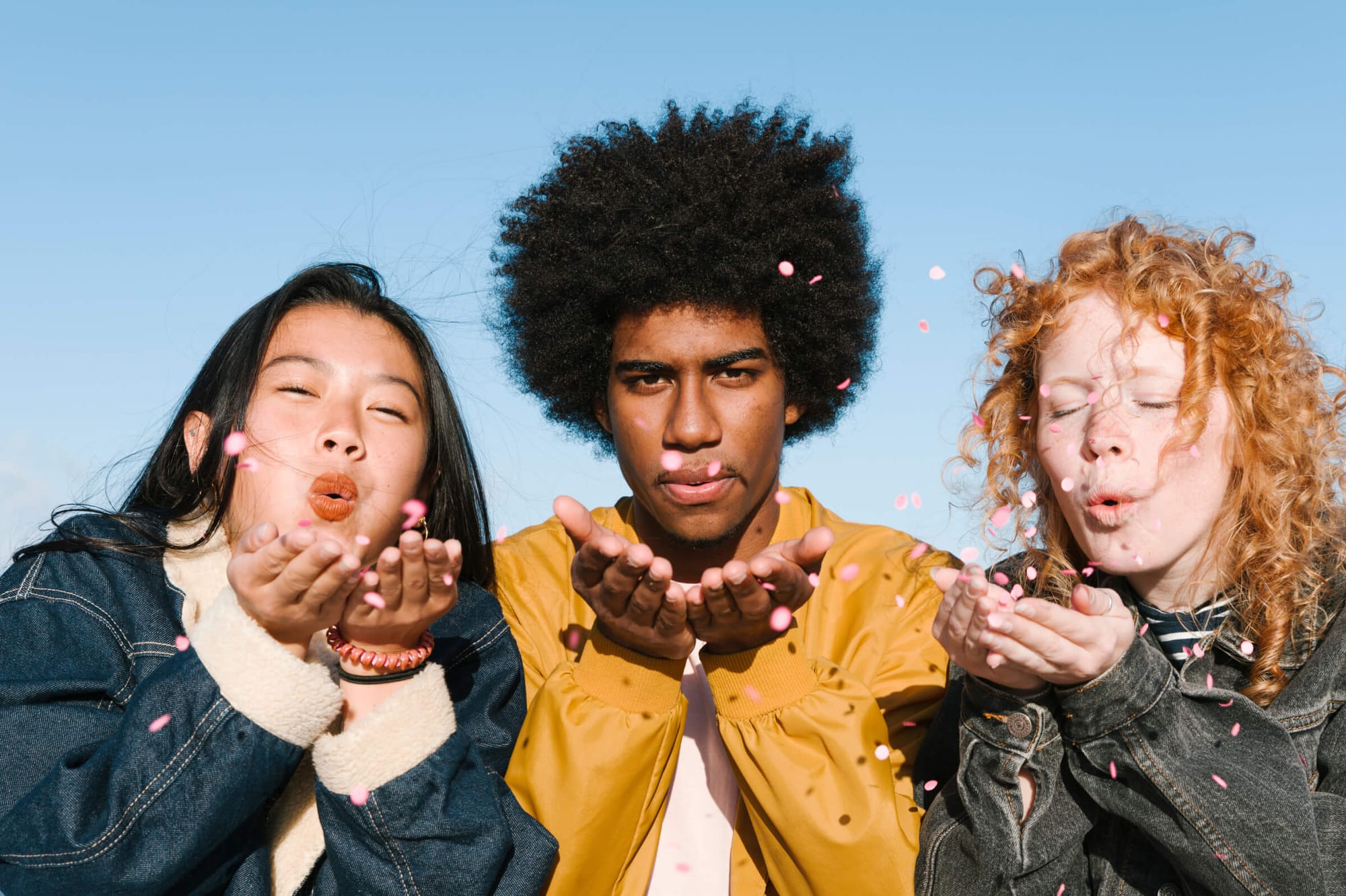 A woman in a denim jacket and a man in yellow blow pink confetti with playful expressions, creating a fun, vibrant scene.