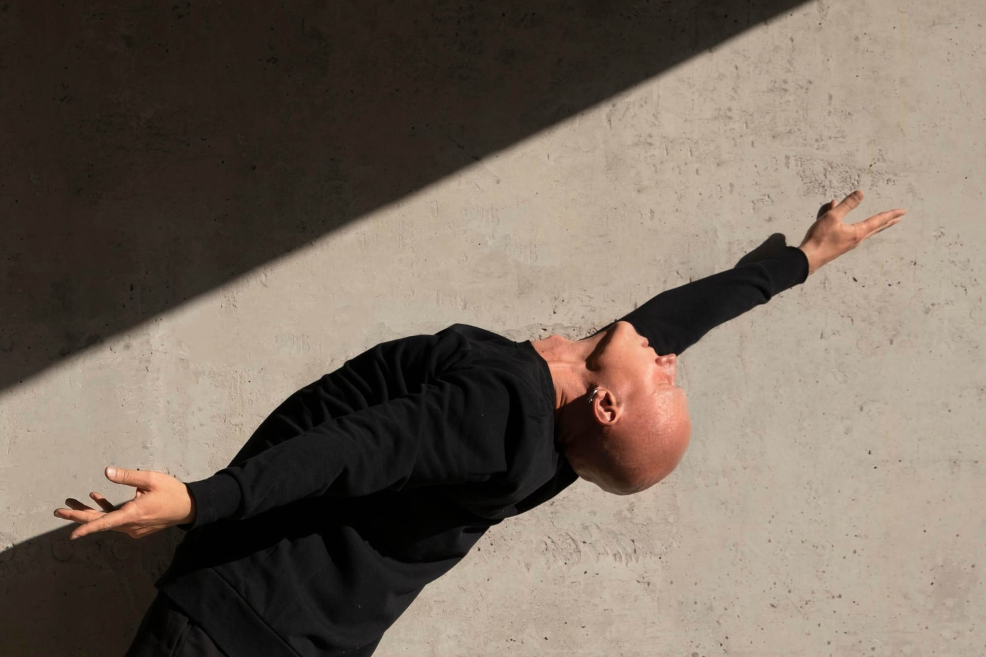 Man in a black outfit striking a dramatic pose against a concrete wall, illuminated by a sharp beam of light.