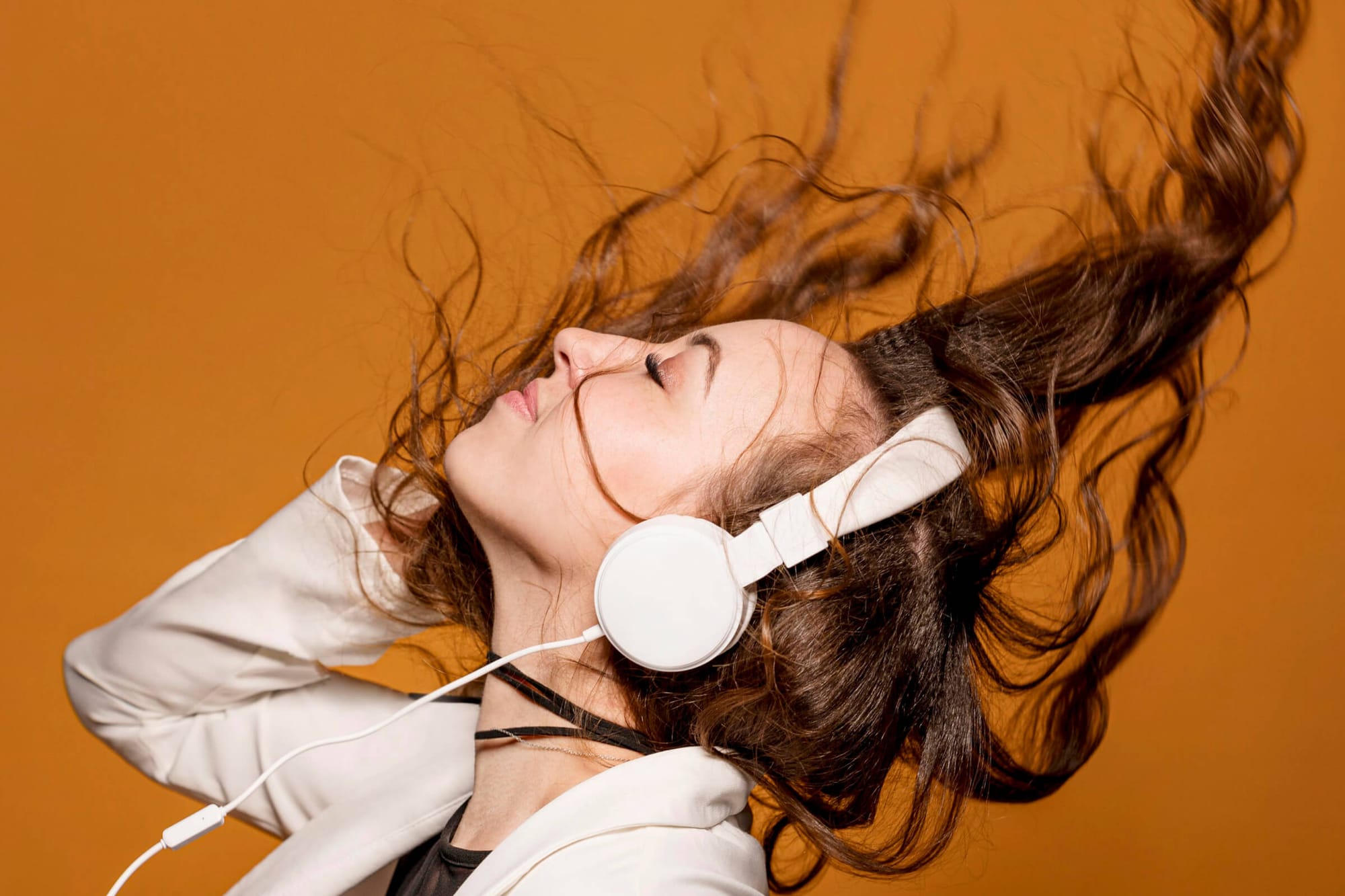 Woman with long hair flipping back while enjoying music on white headphones, set against an orange background.