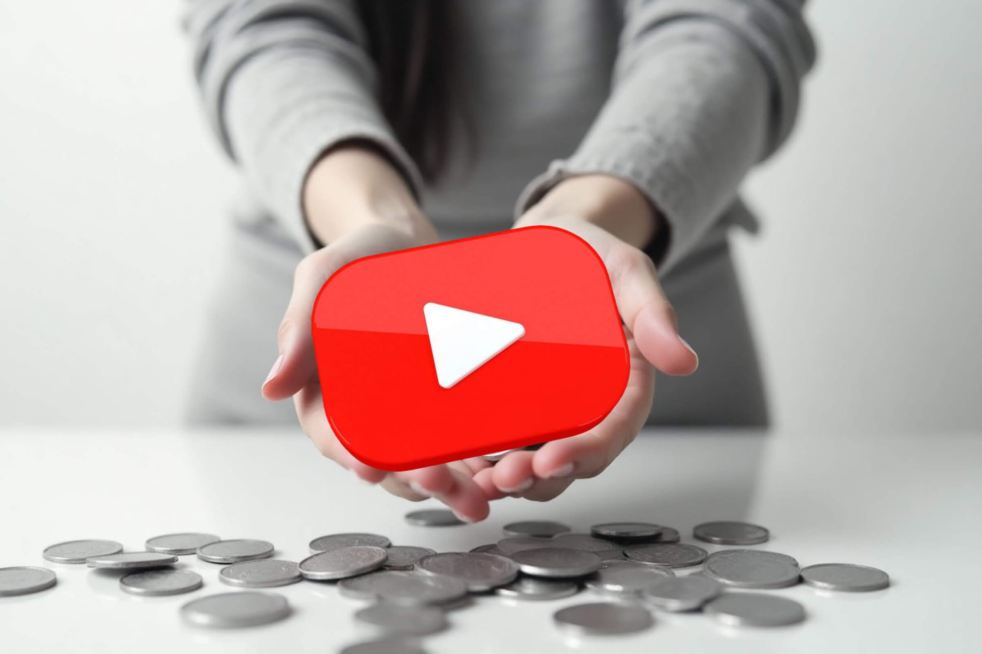 Hands holding a YouTube play button icon over scattered coins.